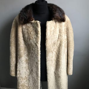 Op de afbeelding: Een lange, cr&egrave;mekleurige schapenvachtjas met een bruine bontkraag.