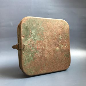 Puede incluir: Una caja cuadrada de metal oxidado con una tapa con bisagras y un cierre de metal. La caja está cubierta de óxido verde y marrón.