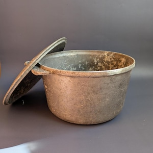May include: A cast iron pot with a lid slightly ajar. The pot is round with a wide opening and a thick rim. The lid has a handle and fits snugly on top. The pot has a dark, aged appearance.