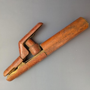 Puede incluir: Una pinza de soldadura vintage, de color marrón rojizo. La pinza tiene un mango tipo palanca y un cuerpo largo y cilíndrico. El metal muestra signos de desgaste y envejecimiento, con un aspecto texturizado. La pinza está diseñada para sujetar electrodos de soldadura.