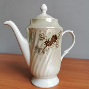 May include: A white ceramic teapot with a swirling design and a brown pine cone pattern. The teapot has a lid and a handle.