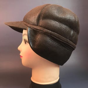 May include: A brown leather hat with ear flaps and a small brim. The hat is displayed on a mannequin head, showcasing its shape and texture. The ear flaps are designed to provide warmth and protection.