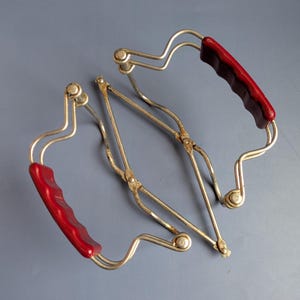 Op de afbeelding: Een paar vintage metalen wecktang met twee gebogen, zilverkleurige draadarmen verbonden door een centraal scharniermechanisme. Elke arm is voorzien van een voorgevormd, felrood kunststof handvat voor een stevige grip tijdens het gebruik.