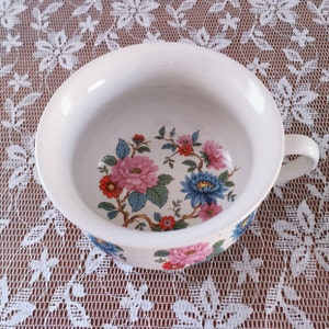 Könnte beinhalten: Weiße Keramikschüssel mit einem Blumendesign mit rosa, blauen und roten Blumen. Die Schüssel hat einen einzelnen Henkel und ist perfekt zum Servieren oder Ausstellen.