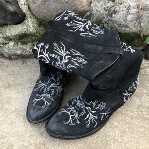 Könnte beinhalten: Ein Paar schwarze Cowboy-Stiefel mit weißen Blumenstickereien. Die Stiefel sind aus Wildleder und haben eine spitze Zehenpartie.