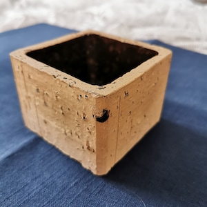 May include: A square, light brown planter made of concrete with a rough, textured surface. The planter has a slightly darker brown interior.