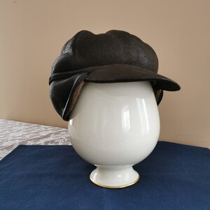 May include: A dark brown leather aviator hat with ear flaps. The hat is displayed on a white, egg-shaped stand with a gold trim. The hat has a textured appearance and a classic design, suitable for cold weather.
