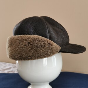 May include: A dark brown, faux leather hat with a rounded crown and a short brim. The hat features a plush, light brown faux fur ear covering. The hat is displayed on a white, decorative pedestal.