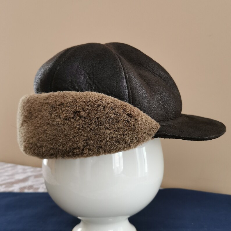 May include: A dark brown, faux leather hat with a rounded crown and a short brim. The hat features a plush, light brown faux fur ear covering. The hat is displayed on a white, decorative pedestal.
