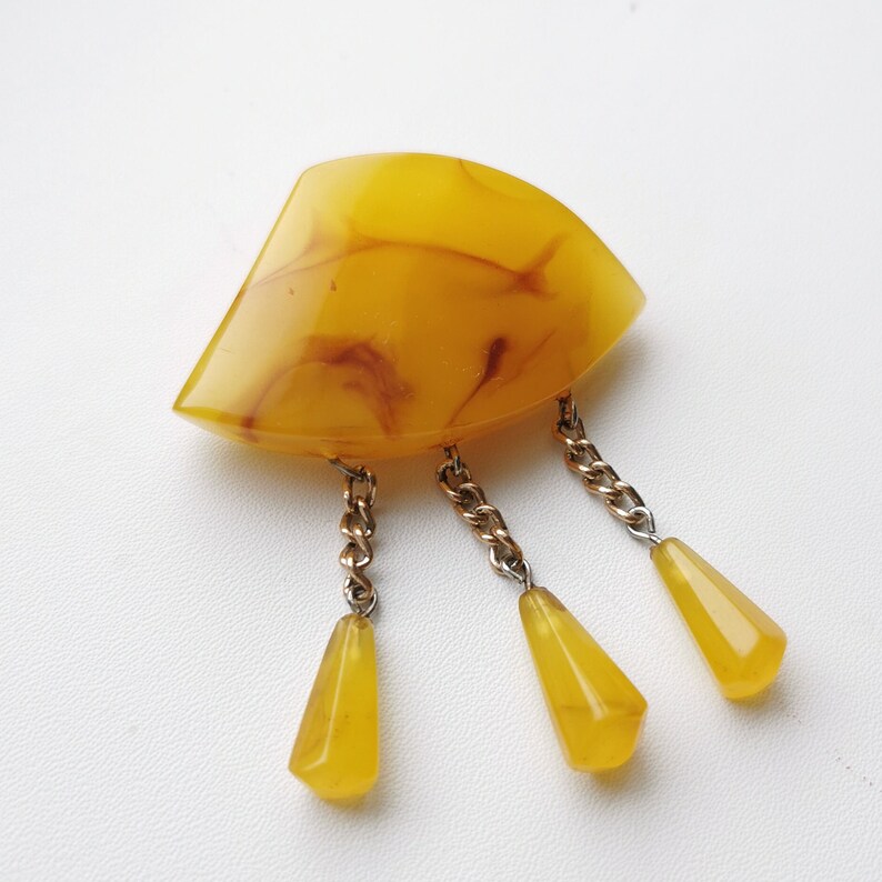 Op de afbeelding: Een gele, gemarmerde, waaiervormige broche met drie hangende gele traanvormige kralen bevestigd aan een gouden ketting.