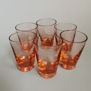 Op de afbeelding: Zes heldere glazen shotglazen met een lichte amberkleurige tint. De glazen zijn in een cirkel op een wit oppervlak gerangschikt.