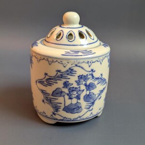 May include: A small, off-white ceramic incense burner with a lid. The lid has several small holes and a rounded knob. The body of the burner is decorated with blue floral and landscape designs. The burner has a rounded shape and sits on three small feet.