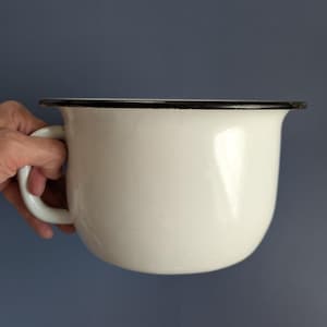 Op de afbeelding: Een witte emaille mok met een zwarte rand en handvat. De mok heeft een afgeronde vorm en een glanzende afwerking. De mok wordt door een hand tegen een blauwe achtergrond gehouden.