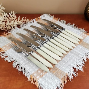 Peut inclure: Un ensemble de 10 couteaux en argent avec des manches de couleur ivoire disposés sur une nappe à carreaux blancs et orange.