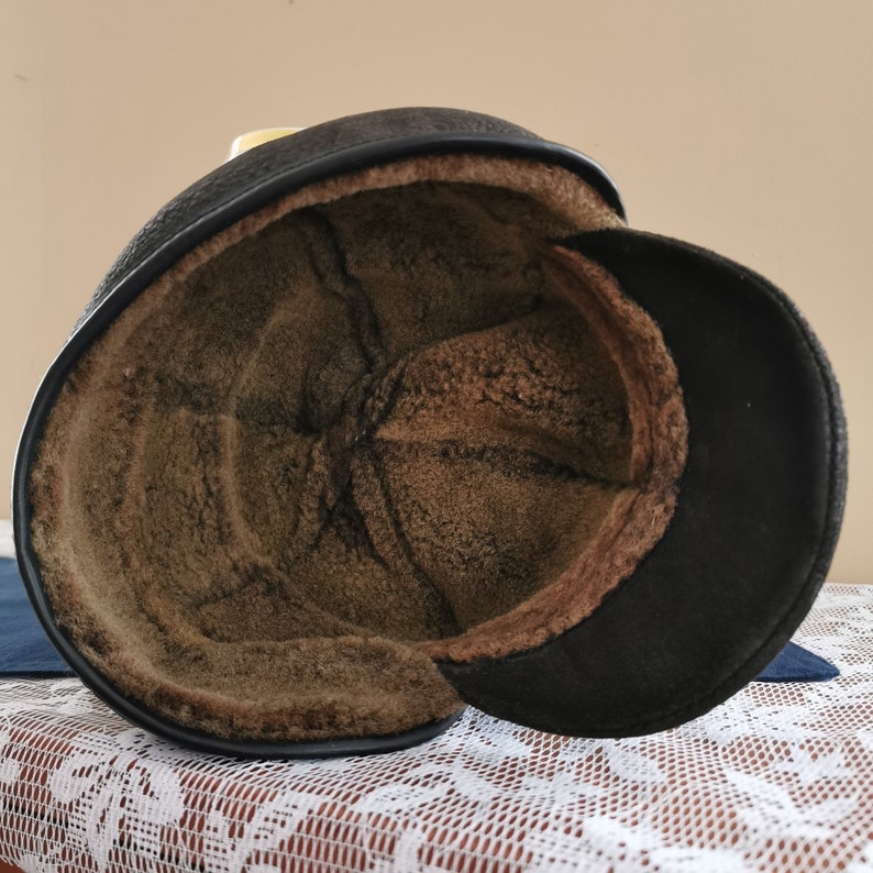 May include: A black leather hat with a brown, fuzzy interior and a small brim. The hat is designed to provide warmth and protection from the elements. The interior is a soft, plush material.
