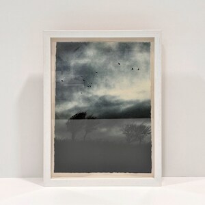 以下が含まれることがあります： ドラマチックな空とシルエットの木々が描かれた風景画の額入りアートプリント。暗く曇り空に鳥が飛び、霧のかかった地平線が描かれています。白い木製フレームに収められています。