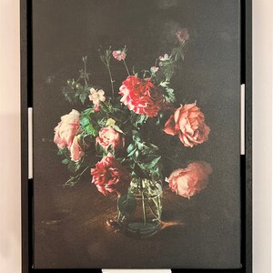 May include: A framed print of a still life featuring a bouquet of pink and red roses in a clear glass vase. The roses are arranged in a vase on a dark wooden table. The background is a dark, textured canvas.