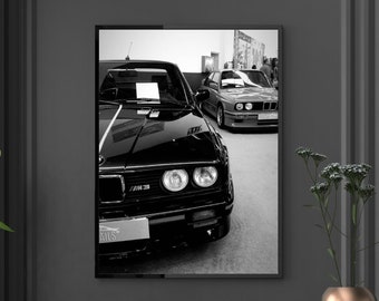 May include: Black and white photograph of a black BMW M3 car. The car is parked in a garage and the front of the car is facing the camera. The car has a black grill and headlights. The car is in focus and the background is blurred.