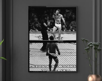 May include: Black and white framed print of a mixed martial arts fight. Two fighters are in the ring, one standing and the other facing him. A referee is in the ring with them. The print is displayed on a dark gray wall above a black dresser.