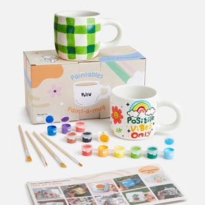 Peut inclure: Un kit de peinture pour mug avec deux mugs en céramique blanche, des pinceaux et une variété de couleurs de peinture. Les mugs sont décorés d'un motif à carreaux vert et blanc et d'un arc-en-ciel avec les mots "Positive Vibes Only".