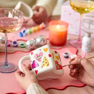 Puede incluir: Una taza de cer&aacute;mica blanca decorada con corazones, ojos y estampados de leopardo. La taza se est&aacute; pintando con un pincel. Una vela naranja encendida, copas de champ&aacute;n y material de pintura est&aacute;n sobre una mesa rosa.