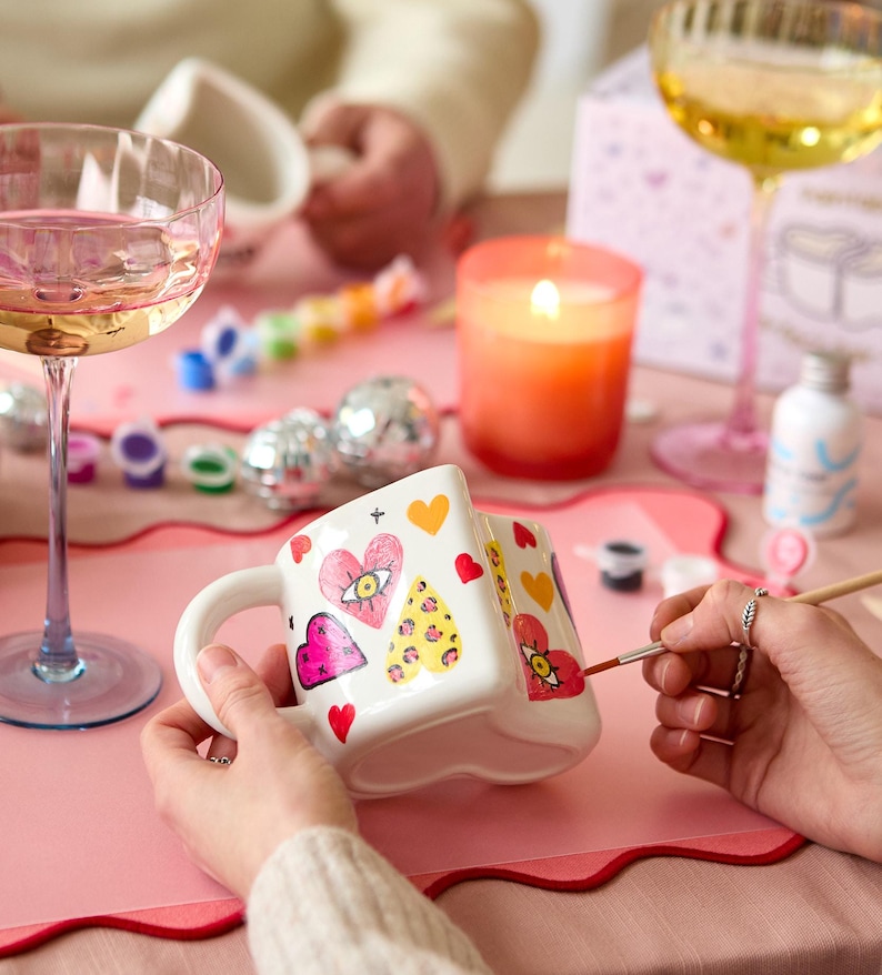 Puede incluir: Una taza de cer&aacute;mica blanca decorada con corazones, ojos y estampados de leopardo. La taza se est&aacute; pintando con un pincel. Una vela naranja encendida, copas de champ&aacute;n y material de pintura est&aacute;n sobre una mesa rosa.