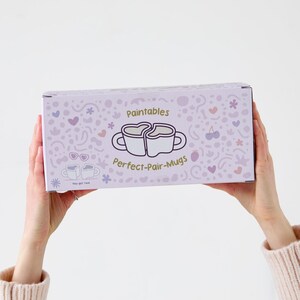 Puede incluir: Una caja rectangular con un fondo lila claro y un patr&oacute;n repetido de corazones, flores y otras formas peque&ntilde;as. La caja presenta el texto "Paintables" y "Perfect-Pair-Mugs" con un gr&aacute;fico de dos tazas formando un coraz&oacute;n.