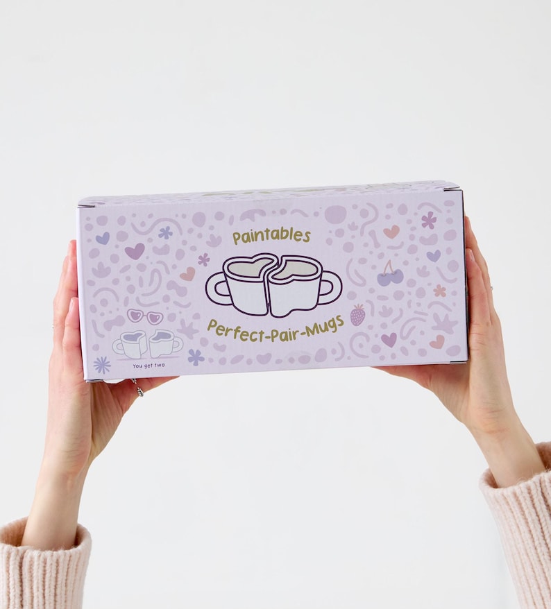 Puede incluir: Una caja rectangular con un fondo lila claro y un patr&oacute;n repetido de corazones, flores y otras formas peque&ntilde;as. La caja presenta el texto "Paintables" y "Perfect-Pair-Mugs" con un gr&aacute;fico de dos tazas formando un coraz&oacute;n.