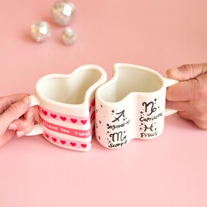Puede incluir: Dos tazas en forma de coraz&oacute;n, una con "I LOVE YOU" y corazones, la otra con los signos del zodiaco Sagitario, Escorpio, Capricornio y Piscis. Las tazas son blancas con detalles rosas, sobre un fondo rosa.