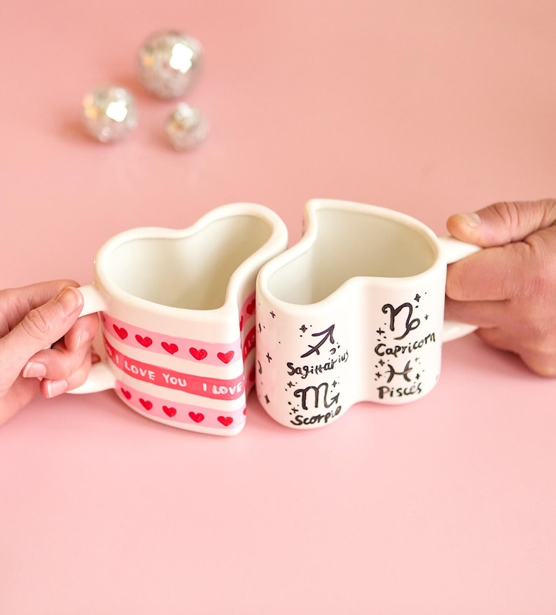 Puede incluir: Dos tazas en forma de coraz&oacute;n, una con "I LOVE YOU" y corazones, la otra con los signos del zodiaco Sagitario, Escorpio, Capricornio y Piscis. Las tazas son blancas con detalles rosas, sobre un fondo rosa.