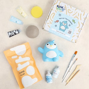 K&ouml;nnte beinhalten: Ein Heim-T&ouml;pferset mit einer Pinguin-f&ouml;rmigen Form, blauer, wei&szlig;er und gelber Farbe, einem Beutel mit Ton und einer durchsichtigen Flasche Glasur. Das Set enth&auml;lt den Text "Patrick The Penguin" und "Home Pottery Kit".