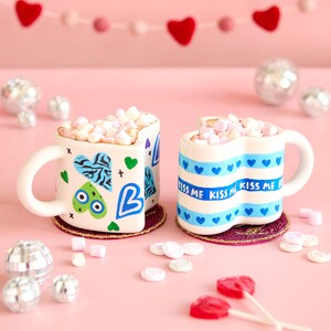 Puede incluir: Dos tazas en forma de coraz&oacute;n llenas de malvaviscos, decoradas con corazones azules y las palabras "Kiss Me". Las tazas est&aacute;n sobre posavasos decorativos, rodeadas de piruletas en forma de coraz&oacute;n y bolas de discoteca, sobre un fondo rosa.