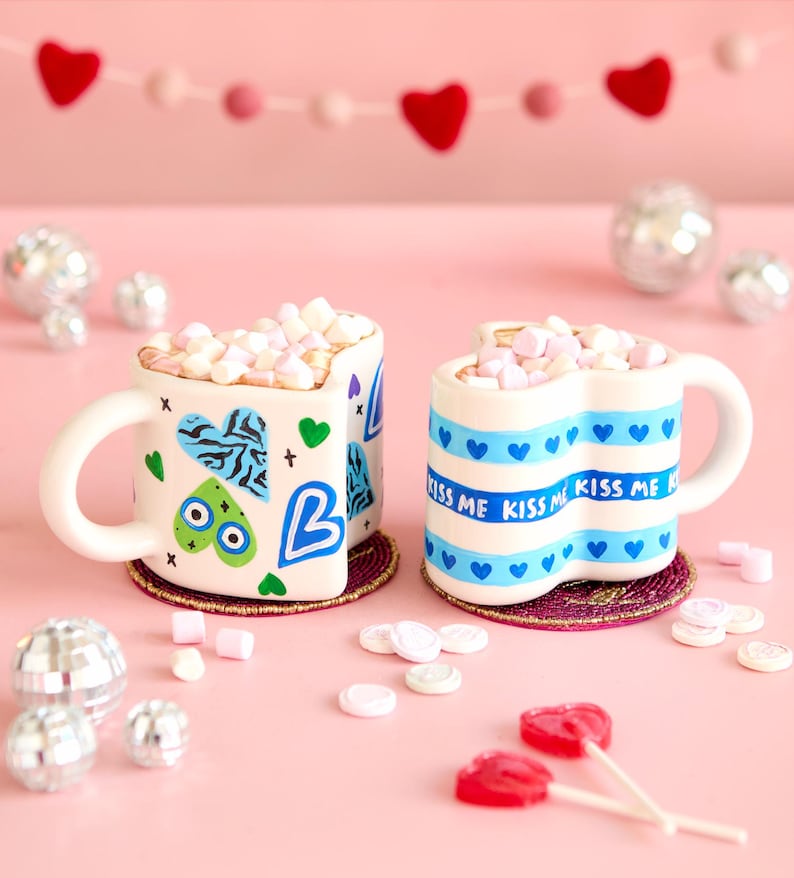 Puede incluir: Dos tazas en forma de coraz&oacute;n llenas de malvaviscos, decoradas con corazones azules y las palabras "Kiss Me". Las tazas est&aacute;n sobre posavasos decorativos, rodeadas de piruletas en forma de coraz&oacute;n y bolas de discoteca, sobre un fondo rosa.