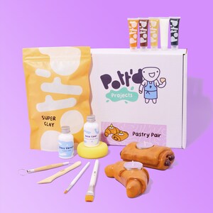 May include: A pottery craft kit with a white box that says "Pottery Projects" and "Pastry Pair". The kit includes a bag of clay, paint, brushes, and two unpainted croissant candle holders.