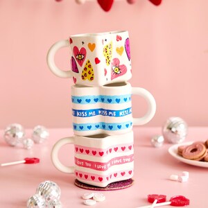 Puede incluir: Tres tazas apiladas en forma de coraz&oacute;n con asas blancas. La taza superior tiene dise&ntilde;os de coraz&oacute;n y ojo. La taza del medio tiene corazones azules y el texto "KISS ME". La taza inferior tiene corazones rosas y el texto "I LOVE YOU".