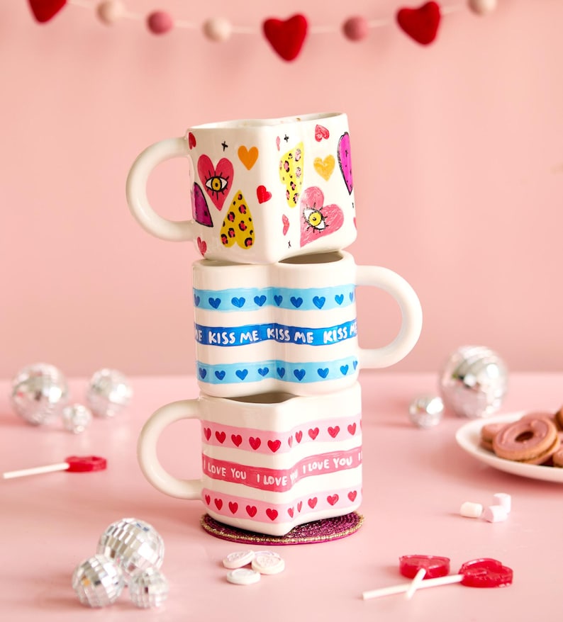 Puede incluir: Tres tazas apiladas en forma de coraz&oacute;n con asas blancas. La taza superior tiene dise&ntilde;os de coraz&oacute;n y ojo. La taza del medio tiene corazones azules y el texto "KISS ME". La taza inferior tiene corazones rosas y el texto "I LOVE YOU".