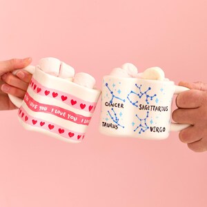 Puede incluir: Dos tazas de cer&aacute;mica blancas llenas de malvaviscos se sostienen sobre un fondo rosa. Una taza tiene una banda rosa con "I LOVE YOU" y corazones rojos. La otra taza presenta constelaciones del zodiaco.