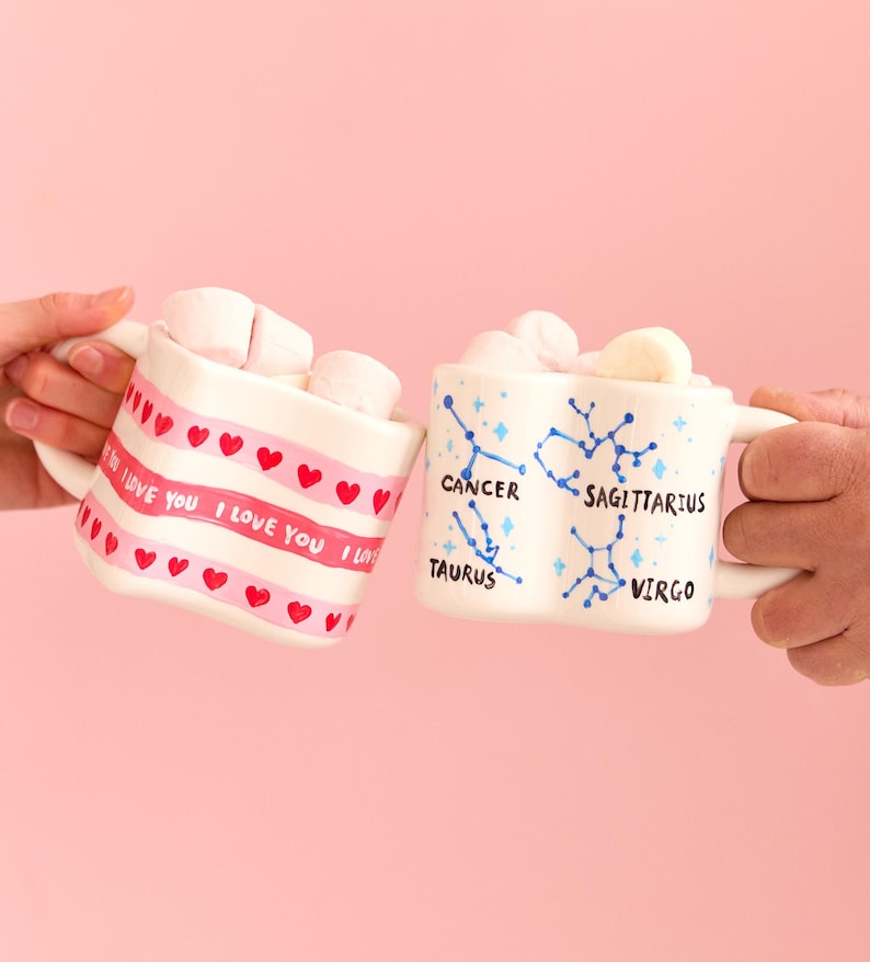 Puede incluir: Dos tazas de cer&aacute;mica blancas llenas de malvaviscos se sostienen sobre un fondo rosa. Una taza tiene una banda rosa con "I LOVE YOU" y corazones rojos. La otra taza presenta constelaciones del zodiaco.
