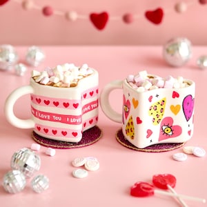 Puede incluir: Dos tazas en forma de coraz&oacute;n llenas de malvaviscos, decoradas con corazones y las palabras "I Love You". Las tazas est&aacute;n sobre posavasos redondos, con un fondo rosa y corazones decorativos.