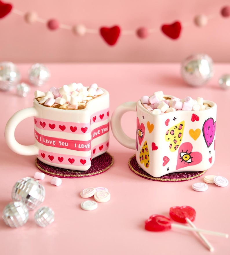 Puede incluir: Dos tazas en forma de coraz&oacute;n llenas de malvaviscos, decoradas con corazones y las palabras "I Love You". Las tazas est&aacute;n sobre posavasos redondos, con un fondo rosa y corazones decorativos.