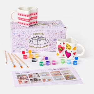May include: A paint-your-own mug kit with two heart-shaped mugs, paint pots, and brushes. One mug has "I LOVE YOU" text and hearts. The other mug has a heart and eye design. The kit includes a box with "Perfect-Pair-Mugs" text.