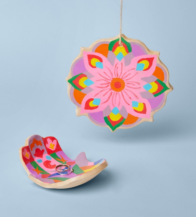 K&ouml;nnte beinhalten: Eine farbenfrohe Keramikschale mit einem rosafarbenen Blumendesign und einem Ring darin. Die Schale ist handf&ouml;rmig und hat ein helles, mehrfarbiges Muster.
