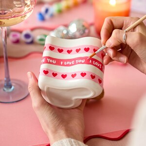 Puede incluir: Una taza de cer&aacute;mica blanca con base en forma de coraz&oacute;n est&aacute; siendo pintada a mano con rayas rosas, corazones rojos y las palabras "I LOVE YOU". La taza est&aacute; sobre una superficie rosa, con una copa de vino y una vela encendida al fondo.