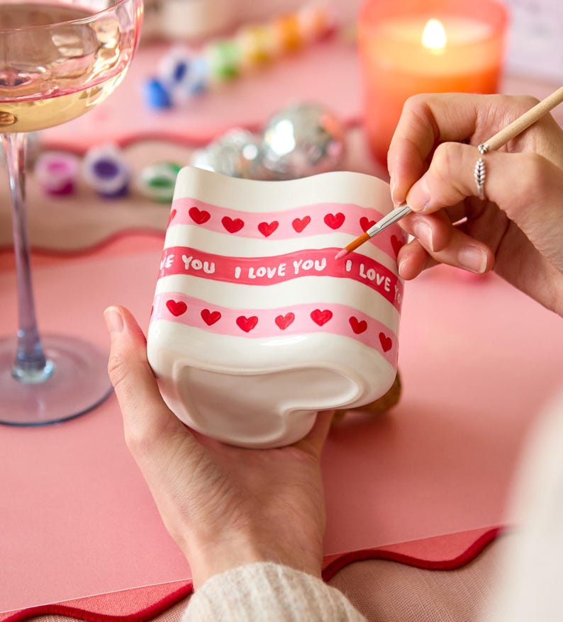Puede incluir: Una taza de cer&aacute;mica blanca con base en forma de coraz&oacute;n est&aacute; siendo pintada a mano con rayas rosas, corazones rojos y las palabras "I LOVE YOU". La taza est&aacute; sobre una superficie rosa, con una copa de vino y una vela encendida al fondo.
