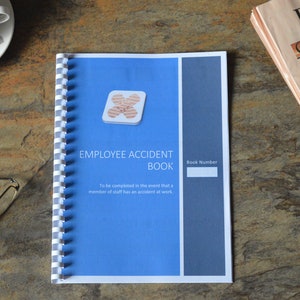 May include: An Employee Accident Book with a blue cover and white spiral binding. The book has the title "EMPLOYEE ACCIDENT BOOK" and a space for a book number. A newspaper, a cup, and glasses are also on the table.