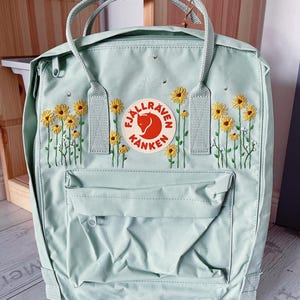Könnte beinhalten: Ein hellminzgrüner Fjällräven Kånken Rucksack mit gestickten gelben Gänseblümchen und grünen Stielen auf der Vorderseite. Der Rucksack verfügt über ein rundes Logo-Emblem mit einem roten Fuchs, eine Reißverschlusstasche vorne und zwei Tragegriffe.