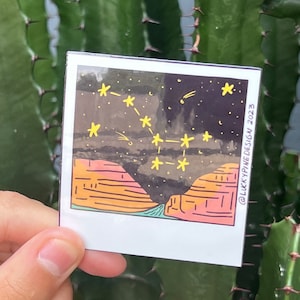 May include: A digital illustration of a starry night sky with a constellation of stars in yellow over a landscape of orange and pink mountains. The image is in a polaroid frame with the text "@LUCKYPINE.DESIGN 2023" in the bottom right corner.