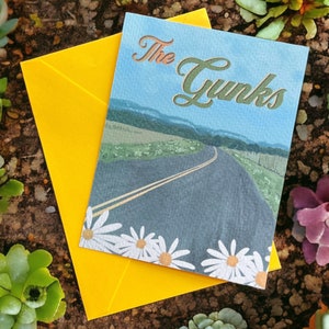 May include: A blue and green card with a road leading through a field of daisies. The text "The Gunks" is written in a cursive font at the top of the card.