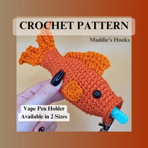Pode incluir: Um porta-caneta vape em forma de peixe laranja de crochê, com olhos pretos e um bocal azul. O texto "CROCHET PATTERN" e "Maddie's Hooks" são visíveis. O texto "Vape Pen Holder Available in 2 Sizes" também é visível.