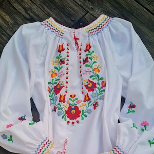 Op de afbeelding: Witte blouse met lange mouwen en kleurrijke bloemenborduurwerk. Het borduurwerk heeft rode, gele, blauwe en groene bloemen. De manchetten en de halslijn zijn afgewerkt met een zigzagpatroon in meerdere kleuren.